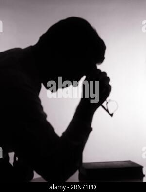 1960ER JAHRE ANONYME SILHOUETTE VON TROUBLED MAN HAND TO STIRN MIT BRILLE MIT BIBEL AUF TISCH BETEN - R19563 HAR001 HARS MÄNNER SPIRITUALITÄT BEUNRUHIGTE B&W SORGEN TRAURIGKEIT HINGABE KOPF UND SCHULTERN RELIGIÖSES BETEN REFLEKTIERENDES DENKEN MEDITATION STIMMUNG REFLEKTIEREND GEDANKEN GEDANKEN VERBINDUNG REFLEKTIEREND BETRACHTEN SIE LOST IN THOUGHT KONZEPTIONELLE UNTERSTÜTZUNG KONTEMPLATIVER ANONYMER TREUER MEDITATIO FAITH MEDITATIVER ELENDER GLAUBE SCHWARZ-WEISS-KONZENTRATION IN ERWÄGUNG ZIEHEN HAR001 ALTMODISCH Stockfoto