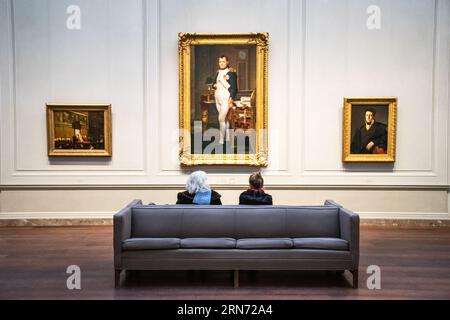 WASHINGTON, DC, USA – zwei Besucher sitzen auf einer Couch in der National Gallery of Art und bewundern Jacques-Louis Davids Meisterwerk „The Kaiser Napoleon in His Study at the Tuileries“ (1812). Das Gemälde, eine berühmte Darstellung von Napoleon Bonaparte, ist in der ständigen Sammlung des Museums ausgestellt und bietet Besuchern ein intimes Erlebnis dieses bedeutenden Kunstwerks der neoklassischen Kunst. Stockfoto