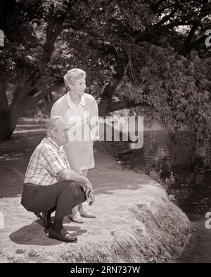 1960ER JAHRE SENIOR EHEPAAR IM PARK MANN KNIEND NEBEN TEICH FRAU STEHEND - S16912 HAR001 HARS EHEMÄNNER GESUNDHEIT NATUR KOPIE RAUM FREUNDSCHAFT IN VOLLER LÄNGE DAMEN PERSONEN MÄNNER RUHESTAND SENIOR MANN SENIOR ERWACHSENER SCHWARZWEISS-PARTNER SENIOR FRAU KNIEND RENTNERTÄTIGKEIT GLÜCK IM ALTER WELLNESS OLDSTERS OLDSTER FREIZEITPARKS FREIZEIT ÄLTESTE NEBEN KOOPERATION ENTSPANNUNG ZWEISAMKEIT FRAUEN SCHWARZ-WEISS KAUKASISCHE ETHNIE HAR001 ALTMODISCH Stockfoto