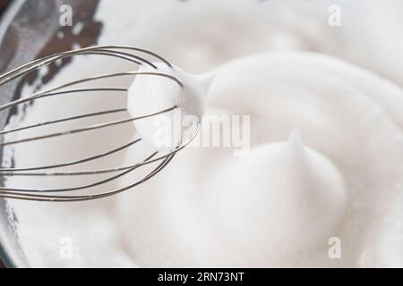 Nahaufnahme von einem Schneebesen und geschlagenem Eiweiß zu steifen Gipfeln, ein Schritt bei der Zubereitung von Frühstücksteig oder zarten Pfannkuchen. Weißer geschlagener Schaum aus Ei W Stockfoto