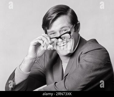 1970ER JAHRE PORTRAIT JUNGER MANN MIT ROLLKRAGENPULLOVER UND SPORTSMANTEL, DER DIE BRILLE ANZIEHT ODER AUSZIEHT, LÄCHELND IN DIE KAMERA SCHAUT - P7979 HAR001 HARS MALES CONFIDENCE BRILLEN B&W AUGENKONTAKT VISION GLÜCK KOPF UND SCHULTERN FRÖHLICHES GUTAUSSEHENDES LÄCHELN FRÖHLICH STILVOLL YUPPIE ODER ANGENEHMES ANZIEHEN ANGENEHMER CHARMANTER KOLLEGIALER ZUSAMMENARBEIT LIEBENSWÜRDIG ANGENEHMER JUNGER ERWACHSENER UND ANSPRECHENDER SCHWARZER UND WEISSE LÄSSIGE KAUKASISCHE ETHNIE HAR001 ALTMODISCHE PREPPY ROLLKRAGEN Stockfoto