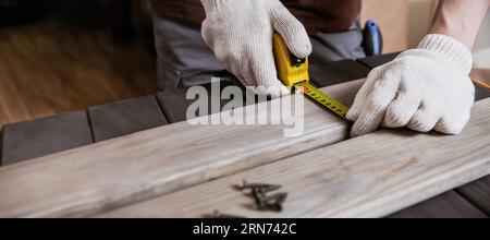 Exakt gemessen. Nahaufnahme eines jungen männlichen Zimmermanns in Handschuhen, der mit gelbem Maßband Messungen auf der Holzdiele anfertigte. Stockfoto