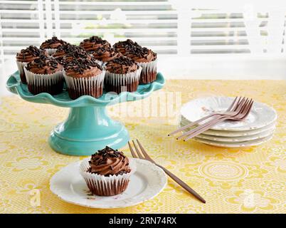 Mini-Schokoladen-Cupcakes auf türkisfarbenem Kakiestand mit kleinen Tellern und Roségold-Gabeln auf gelb gemusterter Tischdecke im horizontalen Format mit Selekti Stockfoto