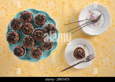 Mini-Schokoladen-Cupcakes auf türkisfarbenem Kakiestand mit kleinen Tellern und Roségold-Gabeln auf gelb gemusterter Tischdecke. Aufnahme von oben in flacher Lay-Komp Stockfoto