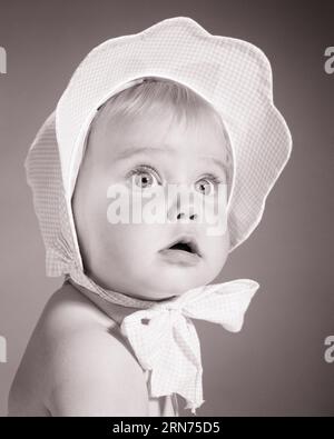 1960ER JAHRE BABY MÄDCHEN MIT UNBESTIMMTEM GESICHTSAUSDRUCK MIT KARIERTER HAUBE GEBUNDEN IN BOGEN UNTER DEM KINN - B22612 HAR001 HARS UND ÜBERPRÜFT ZÖGERLICH UNSICHER STILVOLLE WEITWINKEL ZWEIFELHAFTE MODE WACHSTUM JUNGES BABY MÄDCHEN SCHWARZ-WEISS KAUKASISCHE ETHNIE ZWEIFELHAFT HAR001 UNSCHULD UNSCHULDIG ALTMODISCH UNSICHER Stockfoto