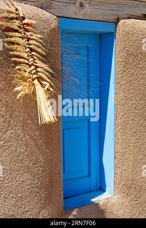 Dekorative Weizenschale hängt am Holzsturz über einem blauen Fensterfenster in lehmziegeln am historischen Ranchos de Taos plaza in New Mexico Stockfoto