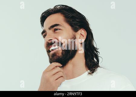 Ein Mann mit Bart und strahlendem Hautton steht in einem Studio und trägt ein perfektes Lächeln im Gesicht. Glücklicher junger Mann, der seine Schönheit und seine männliche Seele umgibt Stockfoto
