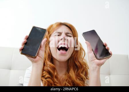 Porträt einer Frau, die schreit, während sie zwei Mobiltelefone und einen Kamerabildschirm zeigt Stockfoto