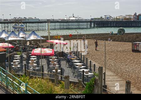 Terrassencafe, Bar und Restaurant am Strand von Brighton in East Sussex, England. Mit Palace Pier im Hintergrund und Menschen am Meer. Stockfoto
