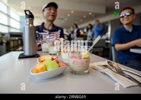 Gäste genießen in der Flughafenlounge ein Frühstück mit Snacks. Mann trägt ein T-Shirt mit der amerikanischen Flagge. Selektiver Fokus auf die Obstschale und das Getränk Stockfoto