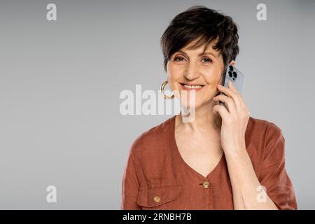 Trendige brünette Geschäftsfrau in brauner Jacke, die auf dem Smartphone redet und auf Grau vor der Kamera lächelt Stockfoto