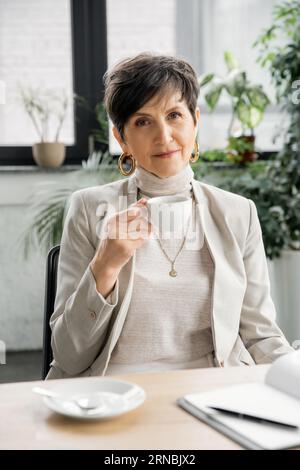 Professionelle Headshot einer erfolgreichen Geschäftsfrau mit Kaffeetasse, die in der Nähe des Notebooks auf die Kamera schaut Stockfoto