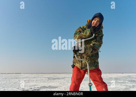 Ein warm gekleideter asiatischer Mann bohrt ein Loch in das Eis auf winterfische Stockfoto