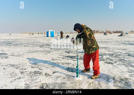 Ein warm gekleideter asiatischer Mann bohrt ein Loch in das Eis auf winterfische Stockfoto