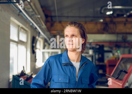 Glücklicher junger Mann in Uniform, der bei Tageslicht in einer verschwommenen Fabrik steht Stockfoto