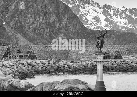 Schwarz-weiß-Foto der Statue der Fischerfrau am Eingang zum Hafen in Svolvaer, getrockneter Kabeljau auf Fish Flakes hinten. Lofoten Island Stockfoto