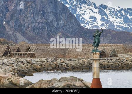 Statue der Fischerfrau am Eingang zum Hafen in Svolvaer, getrocknete Fische auf Fischflocken dahinter. Lofoten-Inseln, Norwegen. 9. Mai 2023 Stockfoto