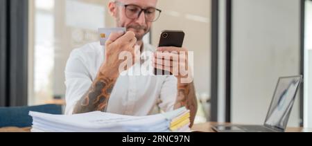 Gestresstes kaukasisches männliches Unternehmen, das Kreditkarte hält und Online-Zahlungen per Smartphone im Büro tätigt Stockfoto