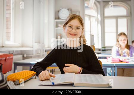 Porträt eines lächelnden Schulmädchens, das eine Brille trägt und mit einem Buch am Schreibtisch im Klassenzimmer sitzt Stockfoto