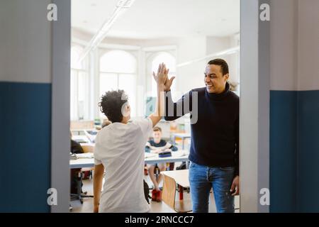 Glücklicher männlicher Lehrer, der dem Schuljungen, der im Klassenzimmer steht, High-Five gibt Stockfoto