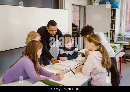 Lehrerin erklärt männlichen und weiblichen Schülern, die im Klassenzimmer auf dem Schreibtisch sitzen Stockfoto