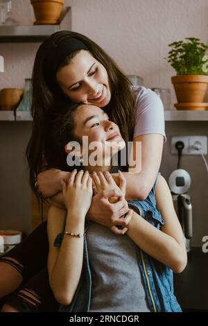 Lächelnde Frau umarmt eine Freundin von hinten, während sie zu Hause in der Küche sitzt Stockfoto