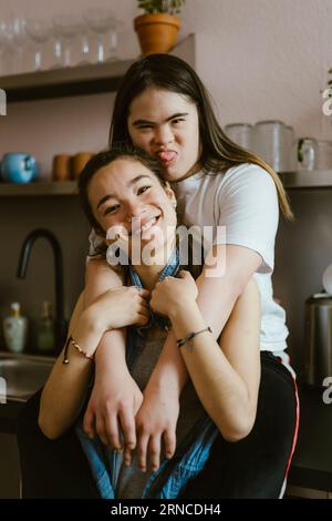 Junge Frau mit Down-Syndrom, die die Schwester von hinten in der Küche umarmt Stockfoto