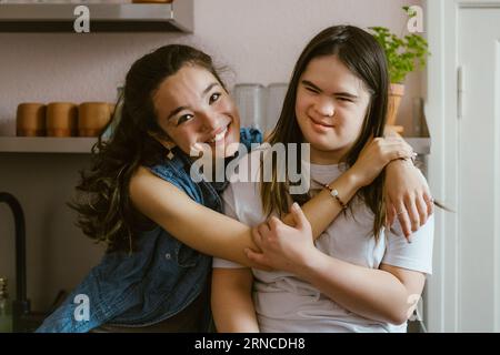 Porträt einer lächelnden jungen Frau, die Schwester mit Down-Syndrom umarmt, in der Küche zu Hause Stockfoto