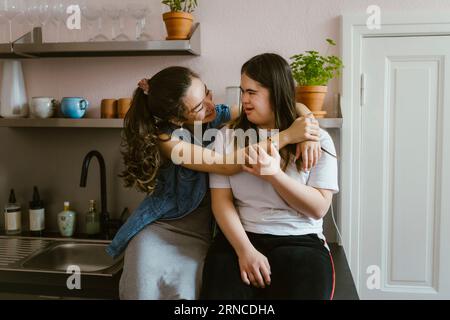 Lächelnde junge Frau umarmt Schwester mit Down-Syndrom in der Küche zu Hause Stockfoto