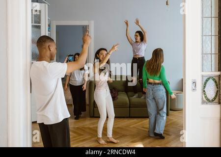 Junger Mann fotografiert Freundinnen, die zu Hause im Wohnzimmer tanzen Stockfoto