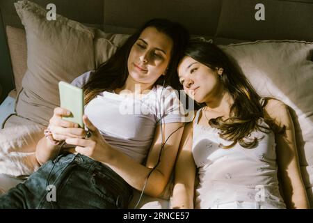 Junge Frauen teilen sich das Smartphone, während sie zu Hause auf dem Bett liegen Stockfoto