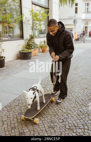 Eine ganze Länge des lächelnden jungen Mannes, der mit Bull Terrier auf dem Skateboard auf dem Bürgersteig läuft Stockfoto