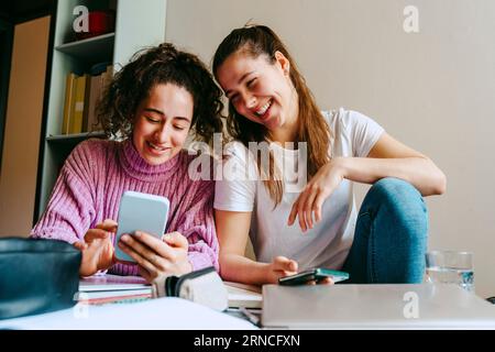Lächelnde junge Frauen teilen sich das Smartphone, während sie zu Hause mit Büchern sitzen Stockfoto