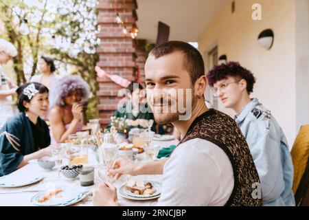 Porträt eines lächelnden schwulen Mannes, der mit LGBTQ-Freunden während einer Dinnerparty im Hinterhof sitzt Stockfoto