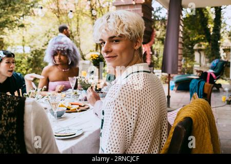 Porträt eines lächelnden schwulen Mannes mit grauen Haaren, der während einer Dinnerparty im Hinterhof unter Freunden sitzt Stockfoto