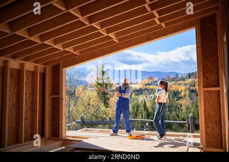 Vater, Mutter und Sohn bauen Holzrahmenhaus. Männlicher Designer in blauen Overalls, der das zukünftige Zuhause zeigt, auf der Terrasse auf der Baustelle. Zimmerei, Familienkonzept. Stockfoto