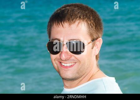 Porträt eines glücklichen jungen Mannes in Sonnenbrille lächelnd auf dem Hintergrund des azurblauen Meeres an einem sonnigen Tag Stockfoto