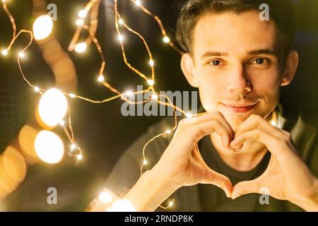 Ein junger Mann in der Nacht, der eine glühende Girlande hält, zeigt eine Herzgeste mit seinen Händen, selektiver Fokus Stockfoto