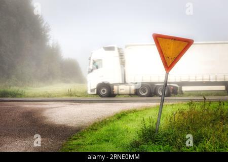 Wegweiser auf einer Landstraße mit weißem Sattelauflieger, der an einem nebligen Tag mit geringer Sicht auf der Autobahn vorbeifährt. Stockfoto