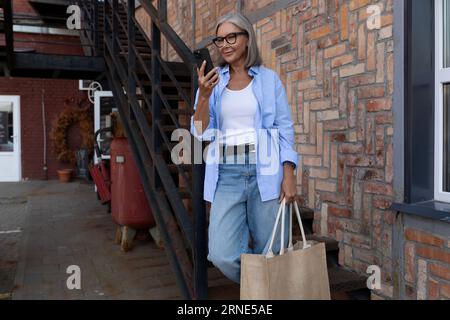 Eine reife grauhaarige Frau im Sommeroutfit spaziert durch die Stadt und chattet am Telefon Stockfoto
