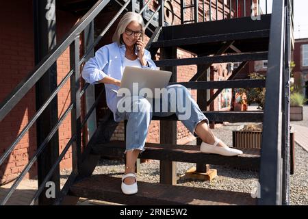 Hübsche, modische Frau mittleren Alters mit grauen Haaren im Sommerstil führt ihr Geschäft mit einem Laptop, während sie auf der Straße sitzt Stockfoto