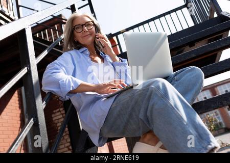 Hübsche, modische Frau mittleren Alters mit grauen Haaren im Sommerstil führt ihr Geschäft mit einem Laptop, während sie auf der Straße sitzt Stockfoto