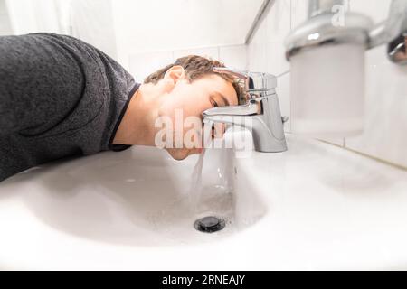 Junger Mann trinkt Leitungswasser mit dem Mund aus dem Waschbecken im Badezimmer mit Lippen und Zunge im fließenden Wasser, das völlig durstig ist und nicht genug Flüssigkeit bekommen kann Stockfoto