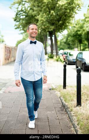 Glücklicher lächelnder Mann mit Fliege und blauem Hemd ist Business-lässig und spaziert am Sommertag auf dem Bürgersteig in der Stadt, während er selbstbewusst und zufrieden ist, wenn er tot ist Stockfoto