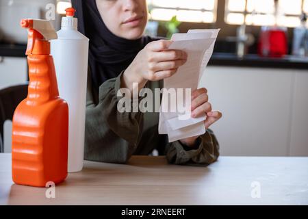 Frau, die in der Küche sitzt und den Kassenbon hält, fühlt sich frustriert wegen der Inflation und der Zunahme des täglichen Bedarfs an Haushaltsmitteln mit Waschmitteln davor Stockfoto