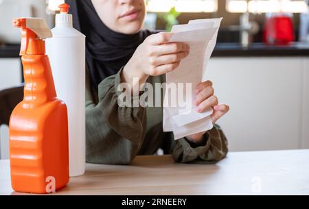 Frau, die in der Küche sitzt und den Kassenbon hält, fühlt sich frustriert wegen der Inflation und der Zunahme des täglichen Bedarfs an Haushaltsmitteln mit Waschmitteln davor Stockfoto