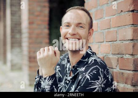 Junger Mann lächelt und schaut auf die Kamera, sitzt bei Tageslicht auf dem Boden neben der roten Ziegelwand und ist glücklich und selbstbewusst Stockfoto