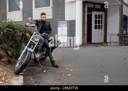 Ein gutaussehender Macho-Biker sitzt auf einem Motorrad und schaut zur Seite, während er in der Altstadt auf der Straße neben einem verlassenen Laden ist Stockfoto