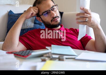 Der Mann ist frustriert von seinen Rechnungen und berechnet sie vor seinem Sofa im Wohnzimmer Stockfoto