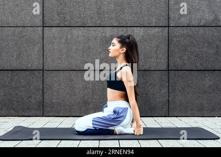 Teenager-Mädchen sitzen in der Stadt draußen auf einer Matte mit Sportbekleidung und geflochtenem Haar, entspannen und meditieren nach dem Yoga-Workout, während sie sich auf Natu konzentrieren Stockfoto